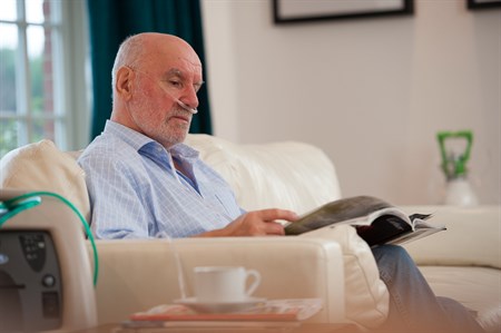 Elderly male patient with oxygen concentrator reading magazine at home