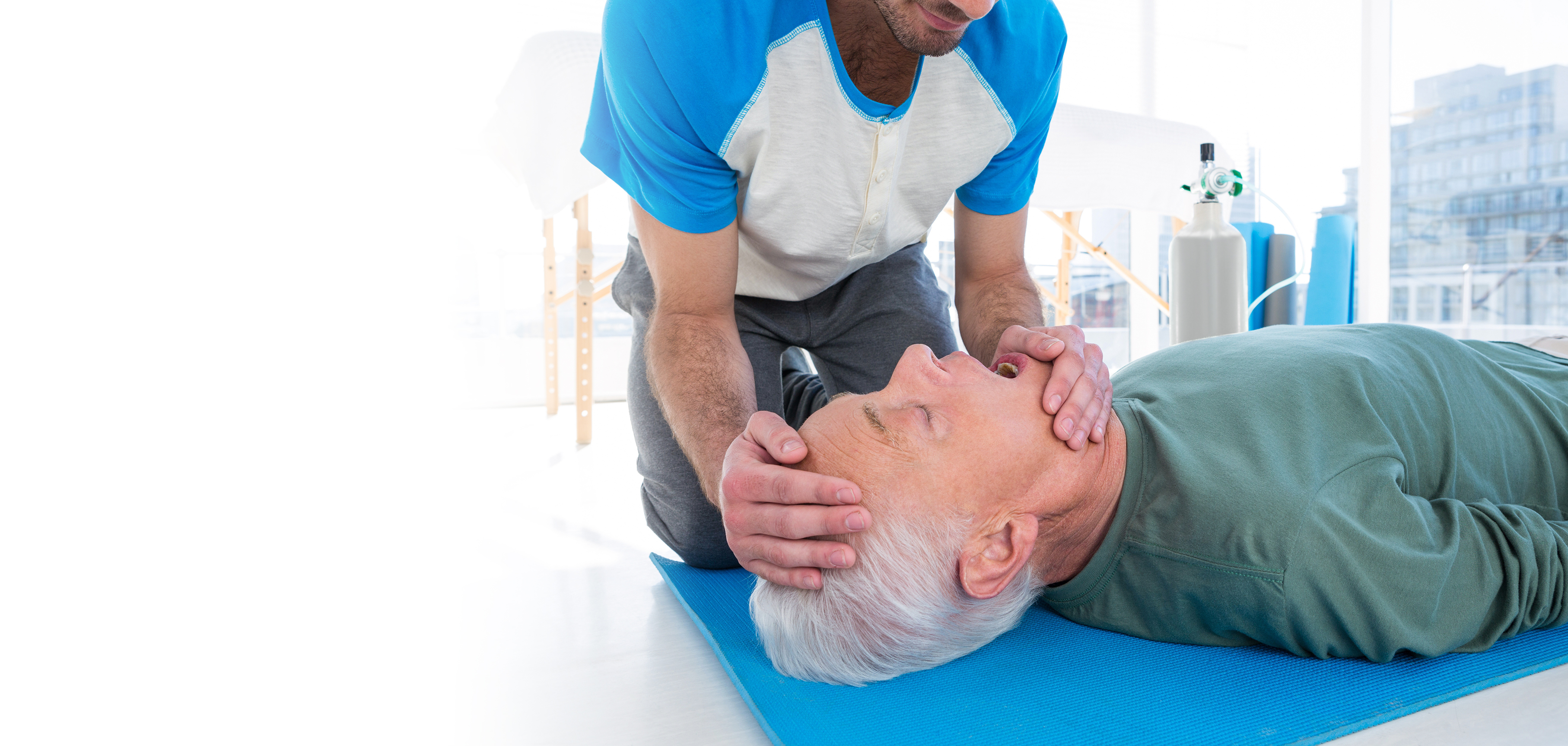 Patient on floor about to receive CPR from man kneeling at his side with emergency oxygen cylinder in the background